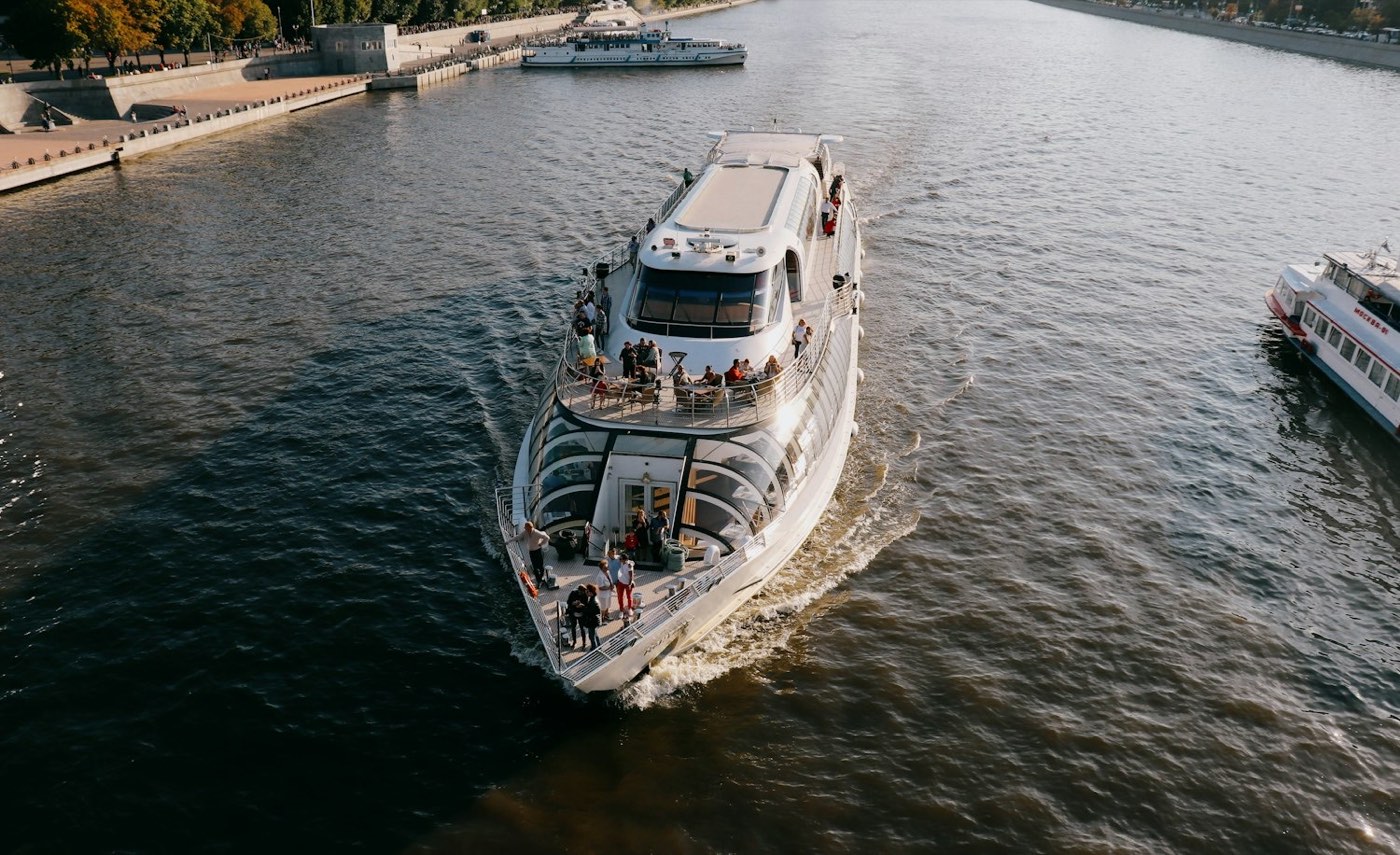 Modernes Fahrgastschiff auf einem Fluss, Gäste stehen an der Reling des offenen Vorderdecks, im Hintergrund eine Uferpromenade